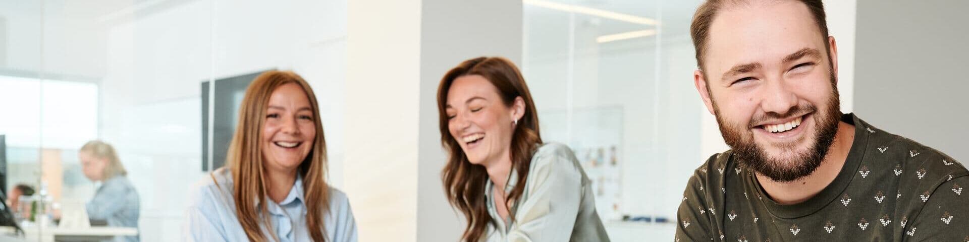 Drei Kolleg:innen lachen und arbeiten gemeinsam an einem hellen Büroarbeitsplatz