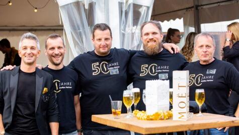 Gruppe von lächelnden Kollegen in schwarzen Shirts mit '50 Jahre Kaffee Partner' Aufdruck feiern gemeinsam, stehen nebeneinander und umarmen sich vor einem Tisch mit Champagner