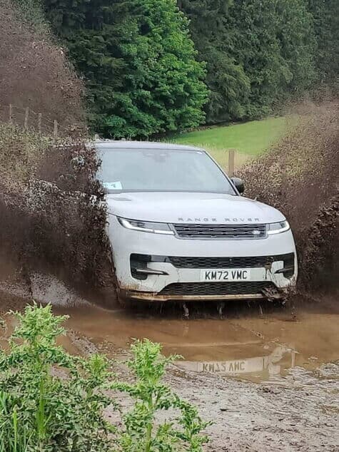 Bei einem Firmen-Teambuilding-Event fahren Mitarbeitende einen weißen Geländewagen durch eine Schlammgrube, dabei spritzt Schlamm auf